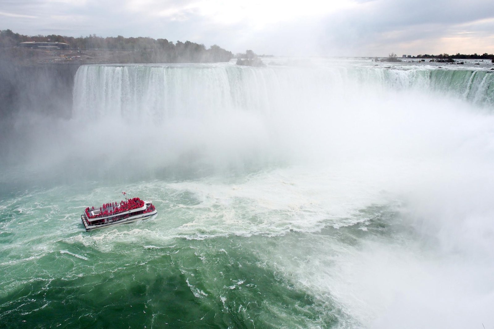 The Horseshoe Falls at Niagara seen from above, with a tour boat crossing the jade-green water below the mist.